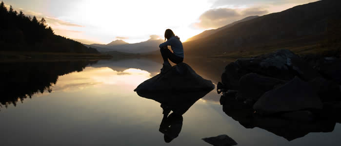 A girl sits on a rock on a lake shore as the sun sets in the distance
